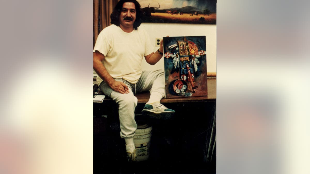 Leonard Peltier sits on a desk while holding a painting toward the camera