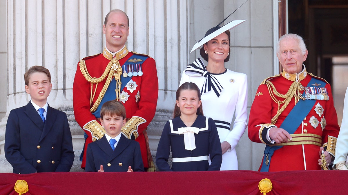 A família real parada na varanda do Palácio de Buckingham olhando para cima em trajes formais.