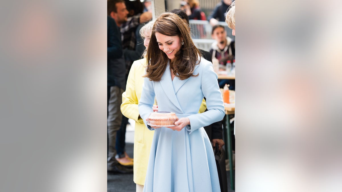 Kate Middleton wearing a pale blue dress smiling and looking down at a cake shes holding.