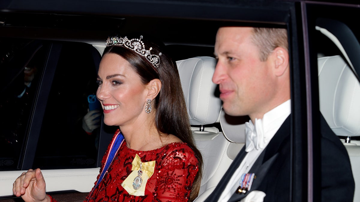 Kate Middleton sorrindo usando um vestido vermelho e uma tiara ao lado do Príncipe William de smoking.