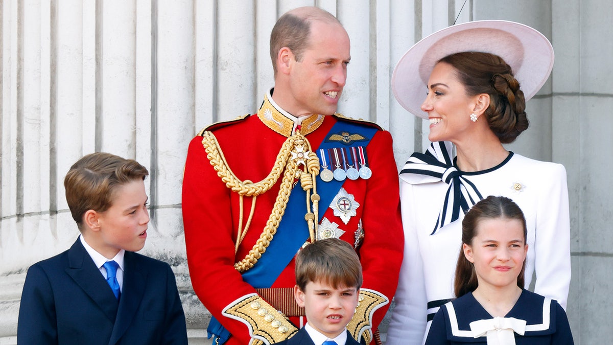 O Príncipe e a Princesa de Gales sorrindo e se admirando na varanda do palácio com seus três filhos.