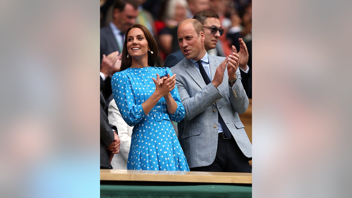 Kate Middleton usando um vestido de bolinhas azul e branco brilhante aplaudindo ao lado do Príncipe William usando um blazer cinza