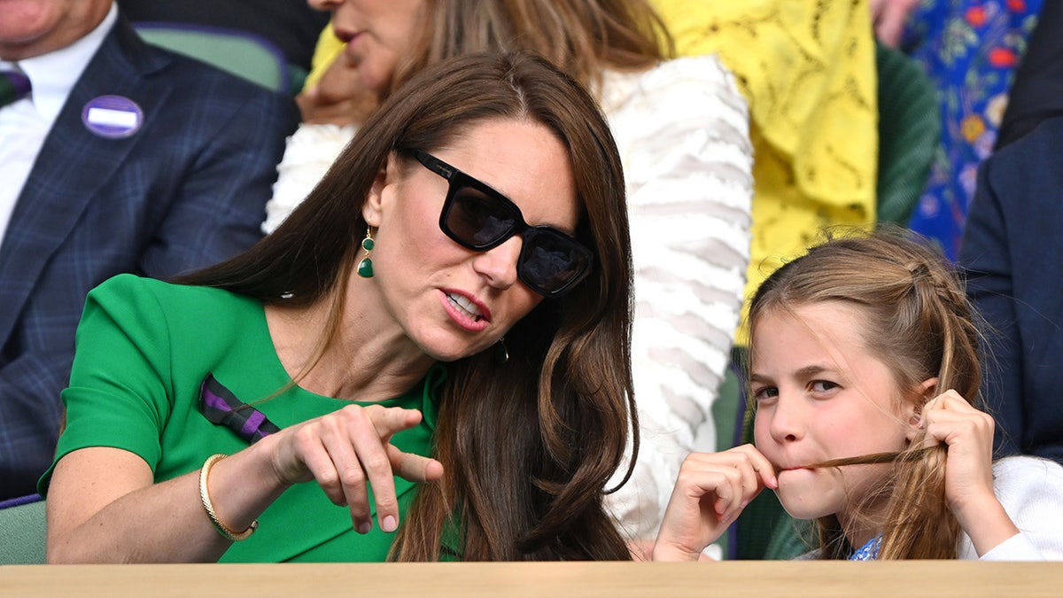 Kate Middleton wearing sunglasses and pointing at something to her daughter Princess Charlotte who has hair in her mouth.