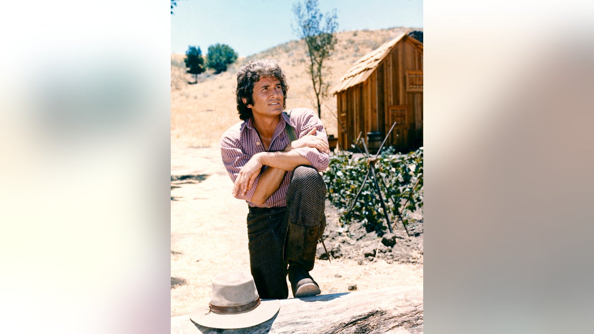 Michael Landon posando con los brazos cruzados y descansando sobre su rodilla con una cabaña de troncos el fondo