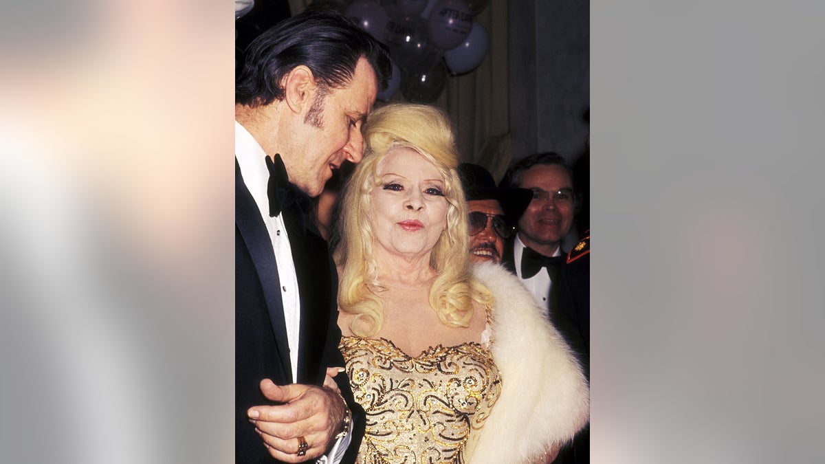 Mae West wears a gold dress on the arm of Paul Novak.