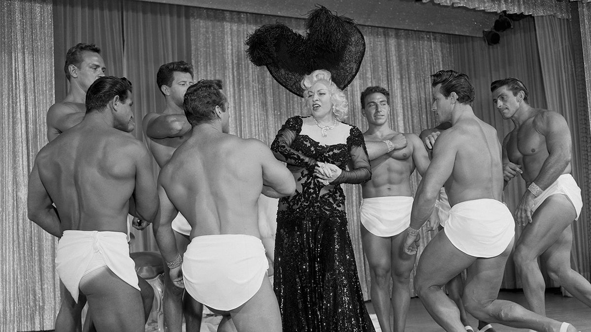 Mae West in a black glamorous gown surrounded by men.