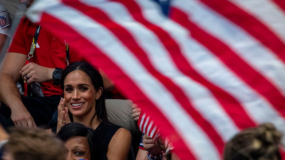 Meghan Markle sitting and smiling as an American flag waves near her.