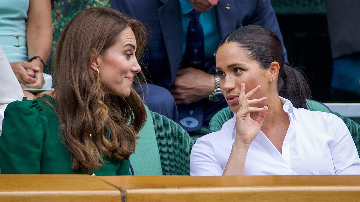 Meghan Markle de blusa branca conversando com Kate Middleton com vestido verde na arquibancada.