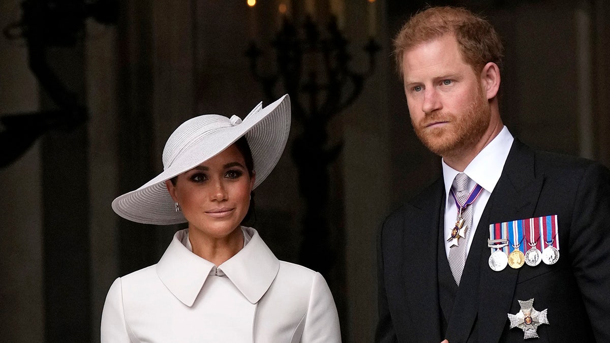 Meghan Markle wearing a white dress with a matching hat standing next to Prince Harry in a dark suit with medals