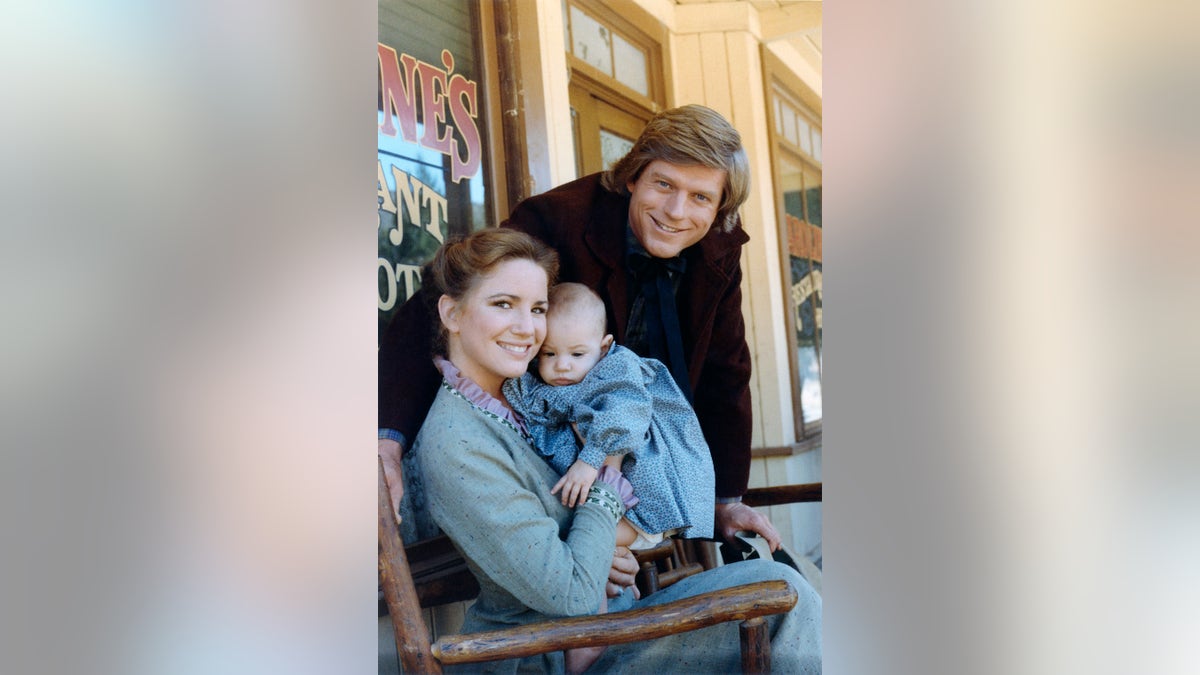 Melissa Gilbert segurando um bebê e sorrindo com Dean Butler posando acima dela também sorrindo.