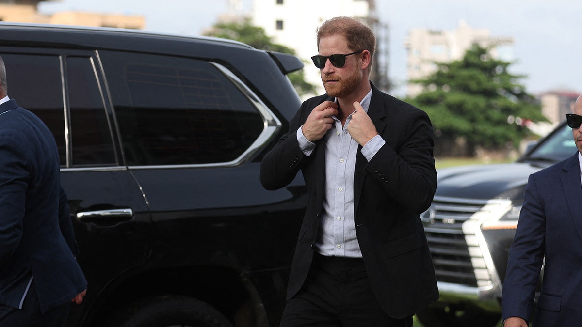 El príncipe Harry con gafas de sol y arreglándose la corbata junto a un coche.