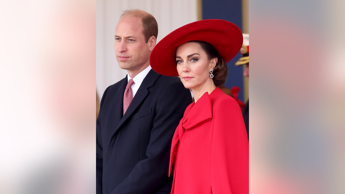 Kate Middleton com uma capa vermelha e um chapéu combinando, ao lado do príncipe William, com um terno escuro e gravata vermelha.