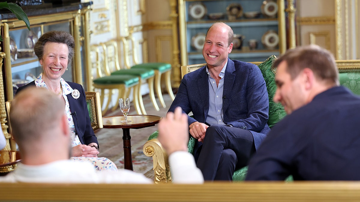Princess Anne smiling and sitting next to Prince William who is also smiling.