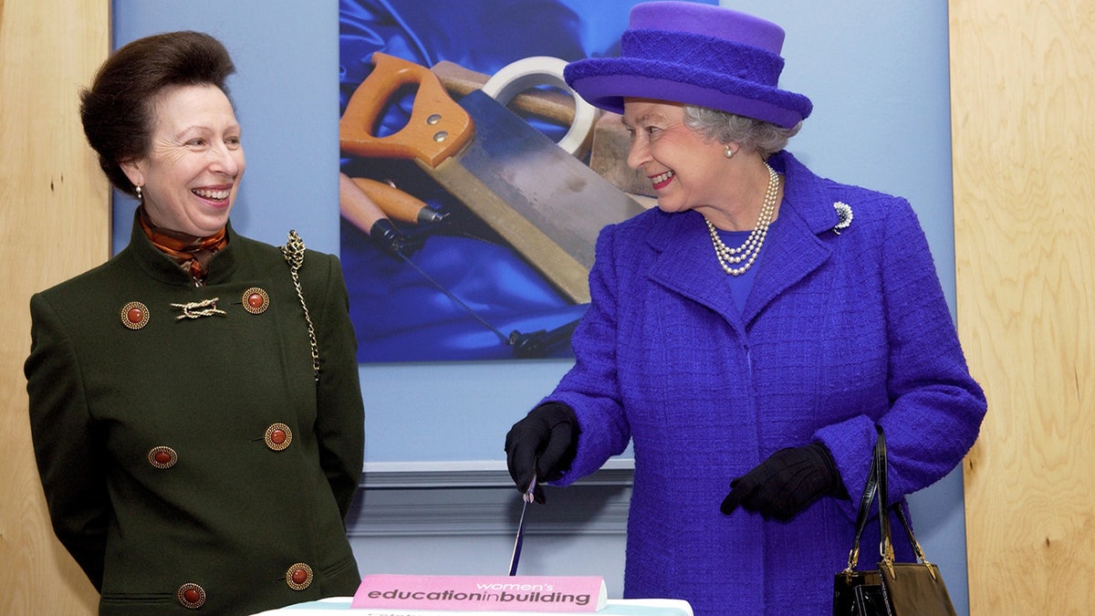 Princess Anne smiling next to Queen Elizabeth who is cutting a cake in a purple suit dress and a matching hat.
