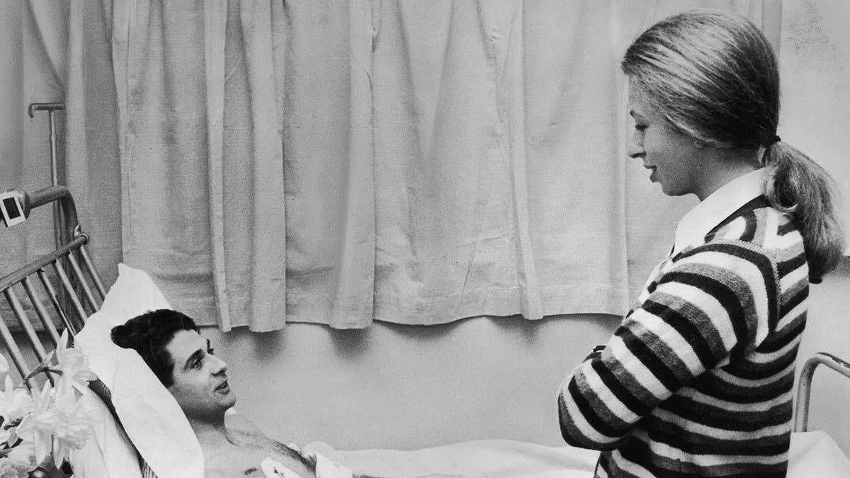 Princess Anne in a striped sweater speaking to a man on a hospital bed.