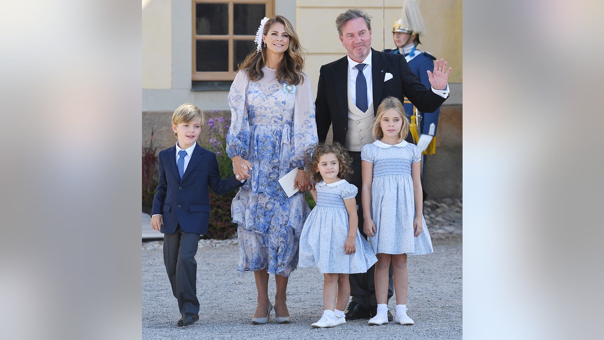 Princess Madeleine of Sweden wearing a white and blue floral dress standing with her husband and their three children all in formal wear