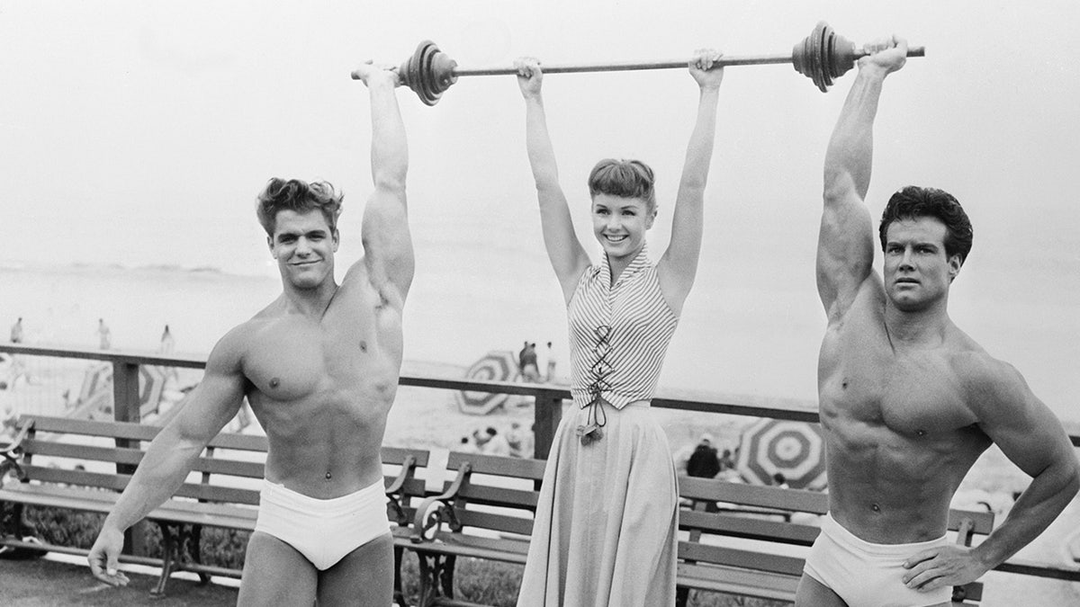 Debbie Reynolds entre dos hombres musculosos levantando pesas.