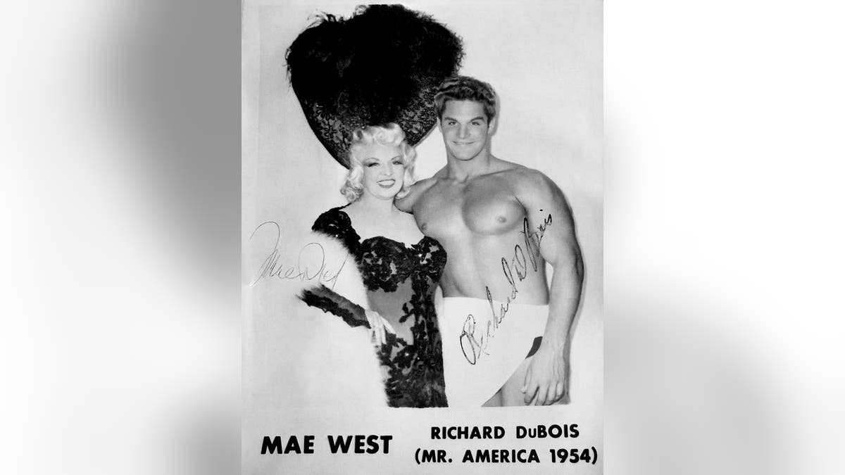 Mae West w kostiumie uśmiecha się i pozuje obok Richarda DuBoisa, który ma ręcznik owinięty wokół talii.