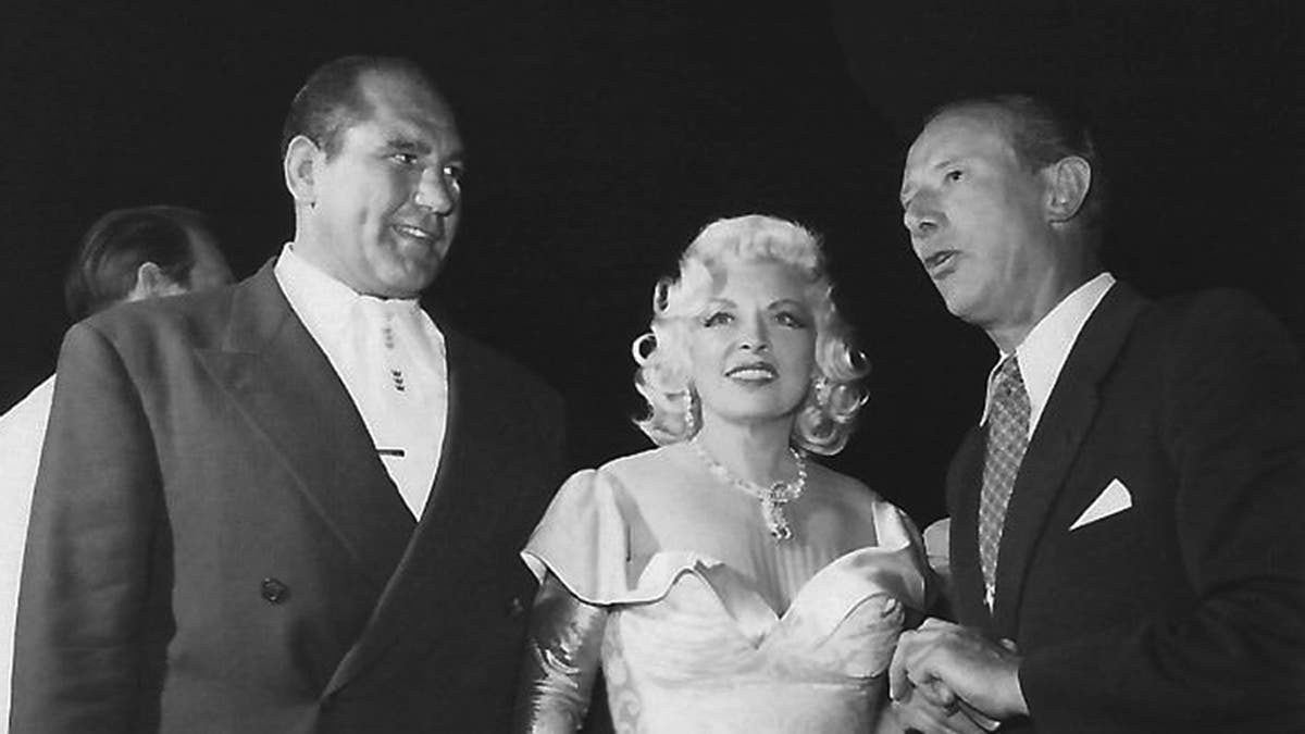 Mae West em um vestido branco glamoroso em pé entre Vincent Lopez e um homem falando