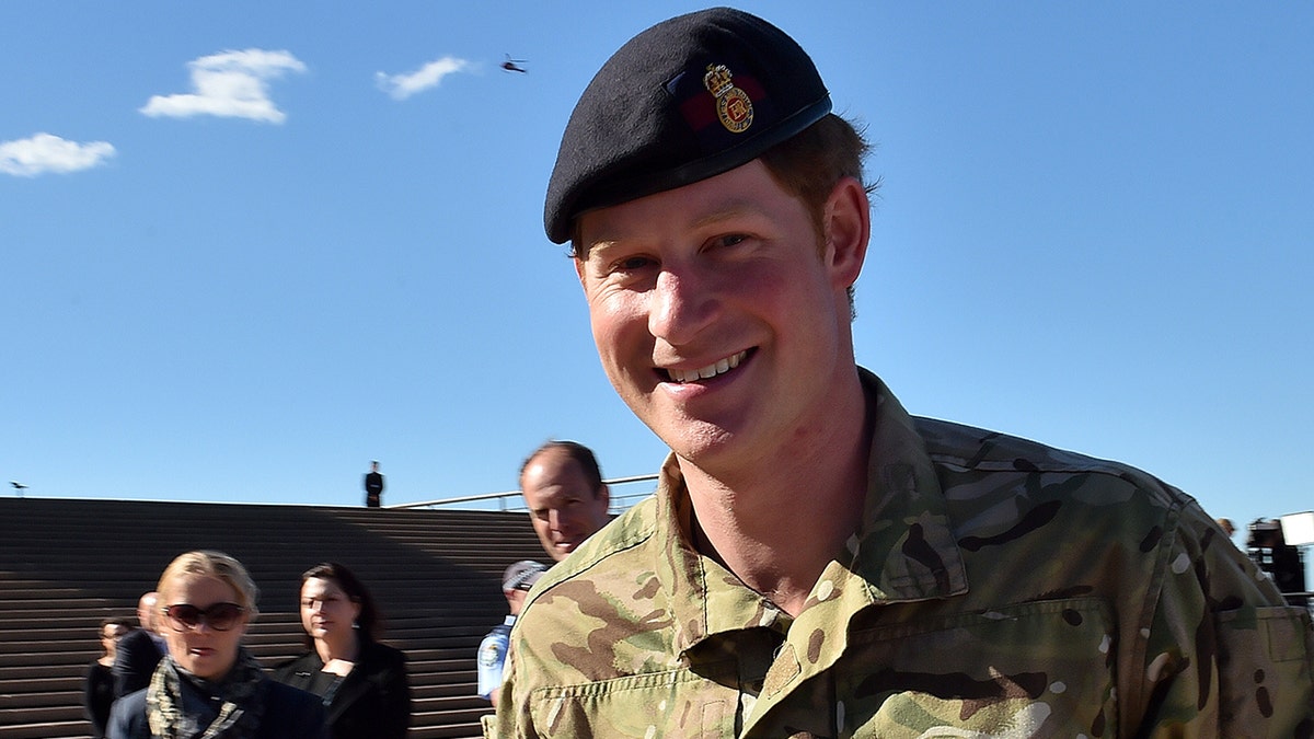 O príncipe Harry sorri com um boné preto e equipamento militar camuflado