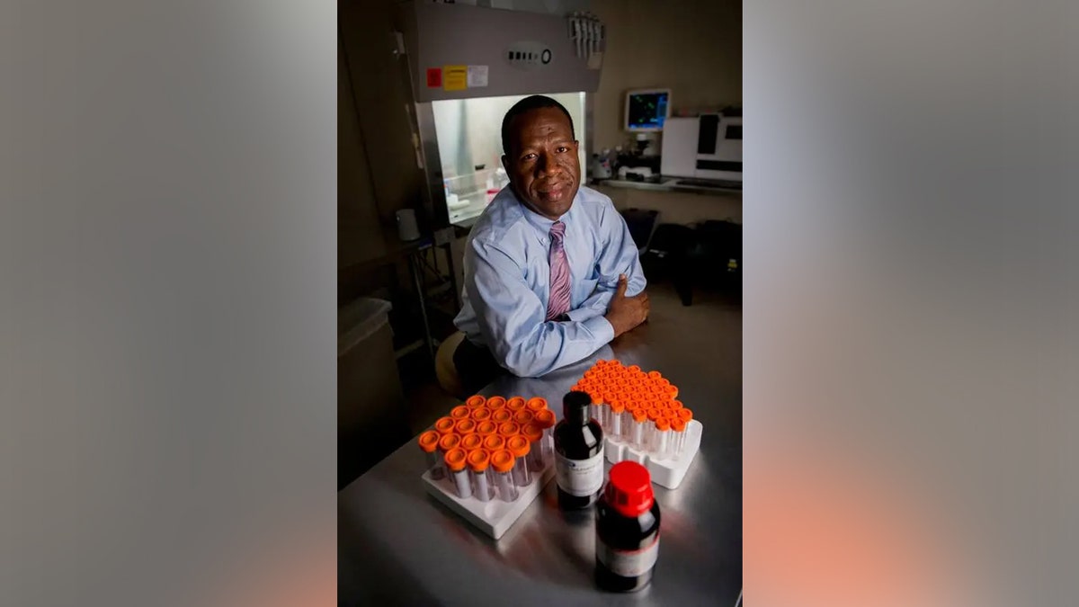 El Dr. Zubair está sentado en su laboratorio, en una mesa detrás de dos bandejas de tubos de ensayo con tapones naranjas.