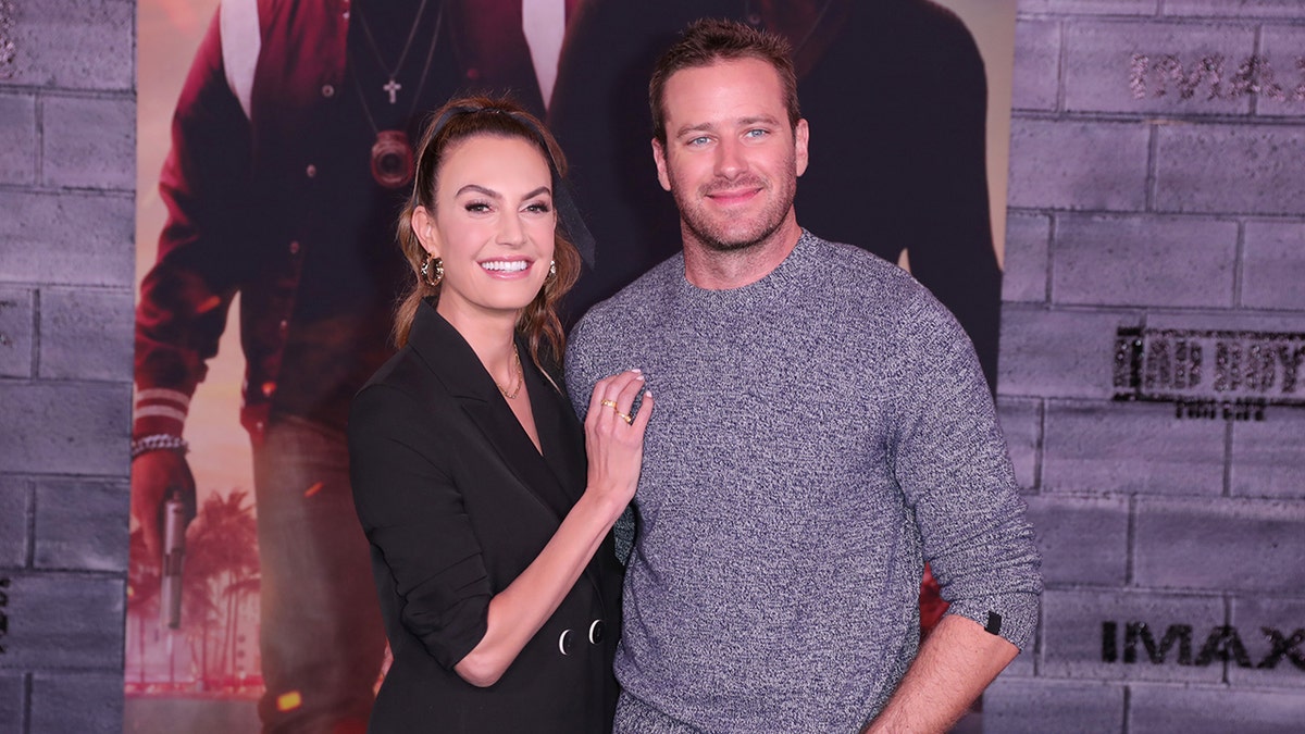 Elizabeth Chambers in black places her hand on Armie Hammer's chest on carpet