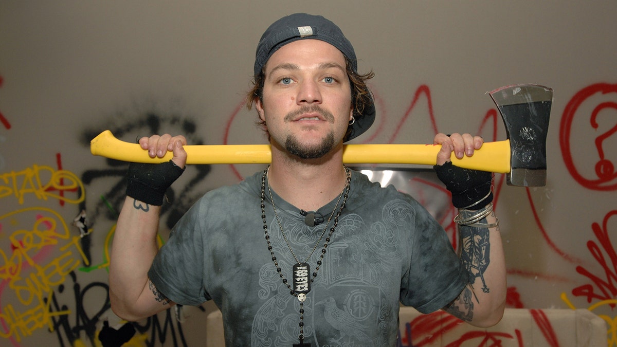 Bam Margera in a grey blue shirt holds an ax behind his back
