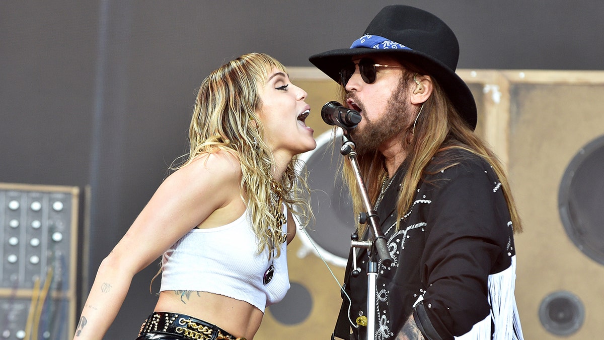 Miley Cyrus in a white crop top sings into one side of the microphone while her dad Billy Ray sings into the other