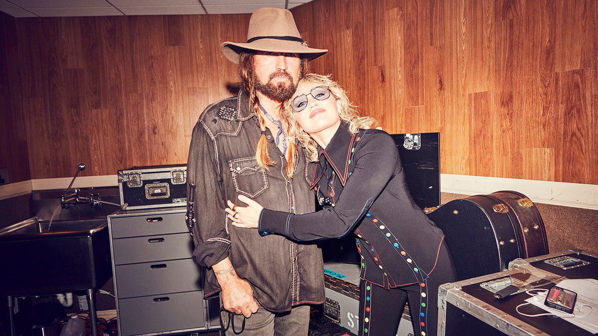 Billy Ray Cyrus in a brown shirt and cowboy hat is hugged by daughter Miley Cyrus in black and clear glasses