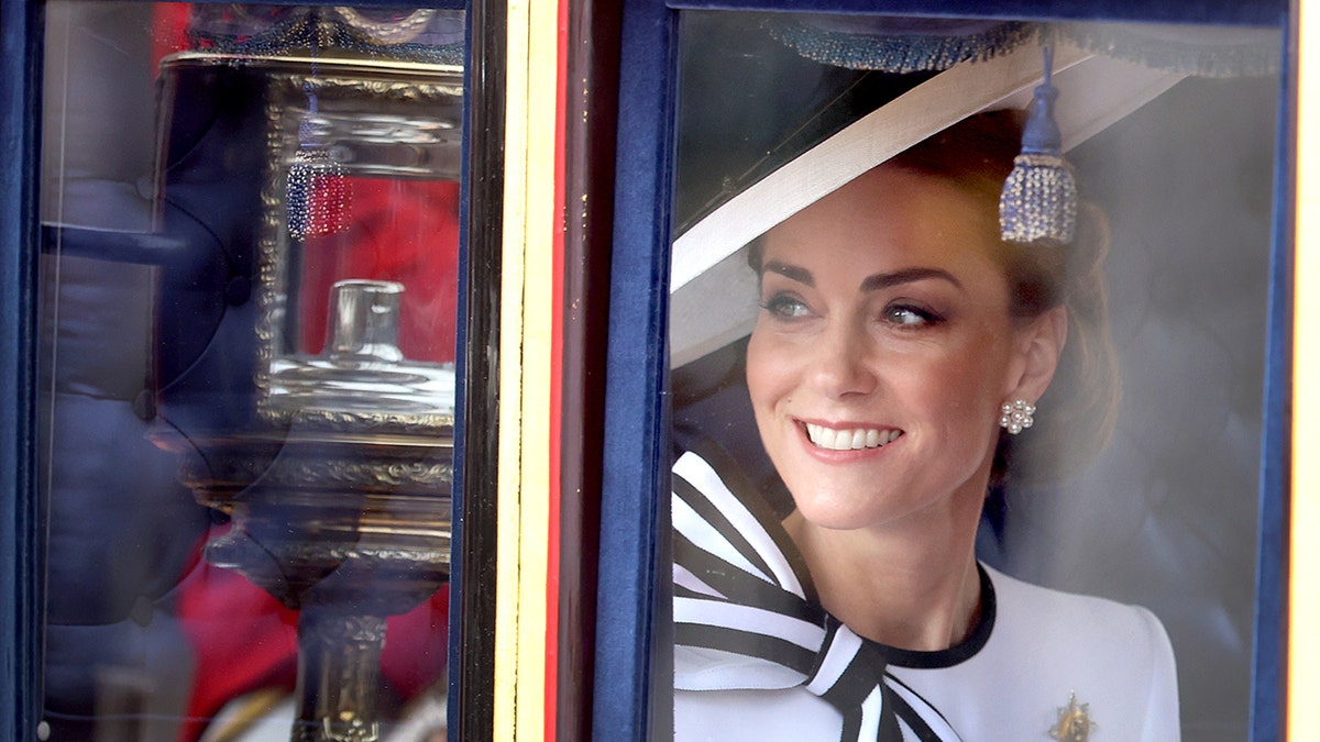 Kate Middleton in a white dress with a striped bow peers out and smiles at crowd during Trooping the Colour arrival
