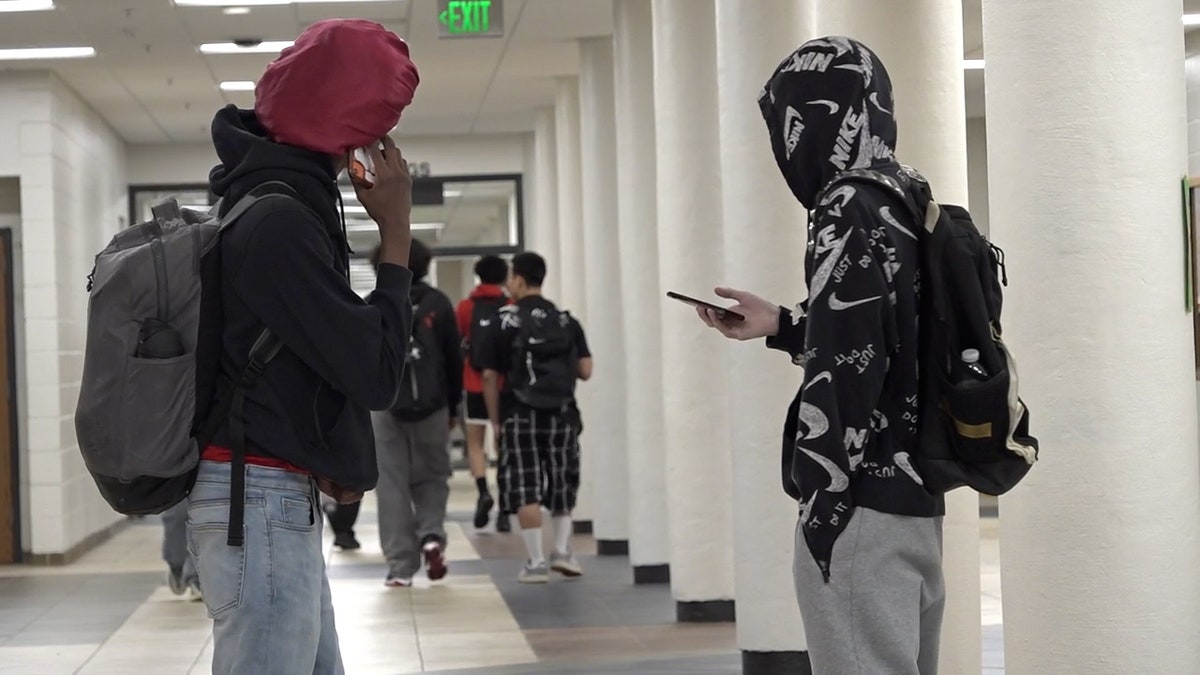 two teenagers talk in a school hallway with one on their phones