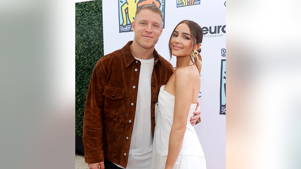Christian McCaffrey in an orange/brown jacket on the carpet with Olivia Culpo in a white dress