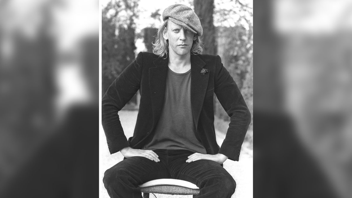 Donald Sutherland sits on a stool wearing a hat and coat in black-and-white image.