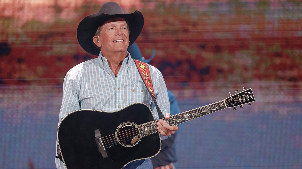 George Strait wears a black cowboy hat and holds a black guitar on stage.