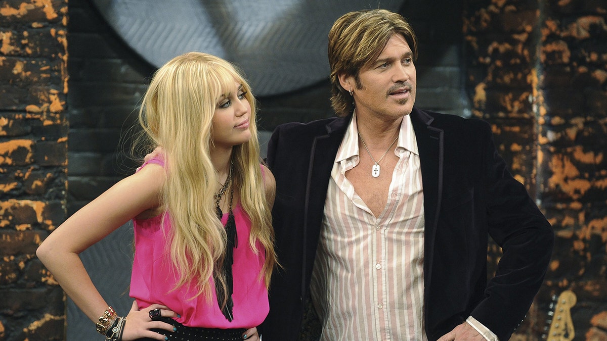 Miley Cyrus como Hannah Montana con peluca rubia y camisa rosa picante con su padre como Robby Stewart