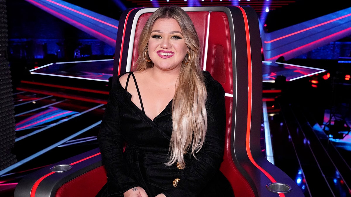 Kelly Clarkson in a black dress sits in a coaches chair on "The Voice"