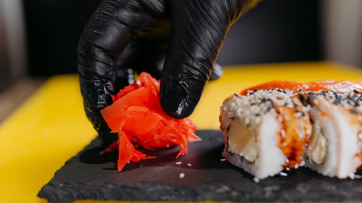 The gloved hand of a sushi chef places ginger on a tray next to some sushi.
