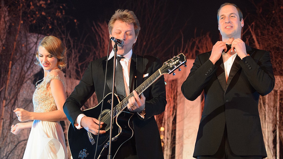 Taylor Swift dança no palco com um vestido branco e dourado enquanto Jon Bon Jovi (Centro) toca violão e o Príncipe William arruma sua gravata borboleta