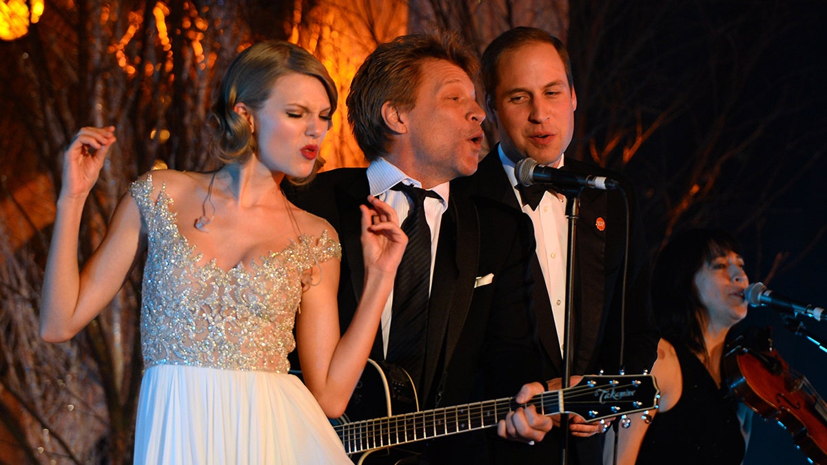 Taylor Swift em um vestido branco e dourado levanta os braços enquanto se apresenta no palco com Jon Bon Jovi de terno preto e gravata e o Príncipe William de smoking