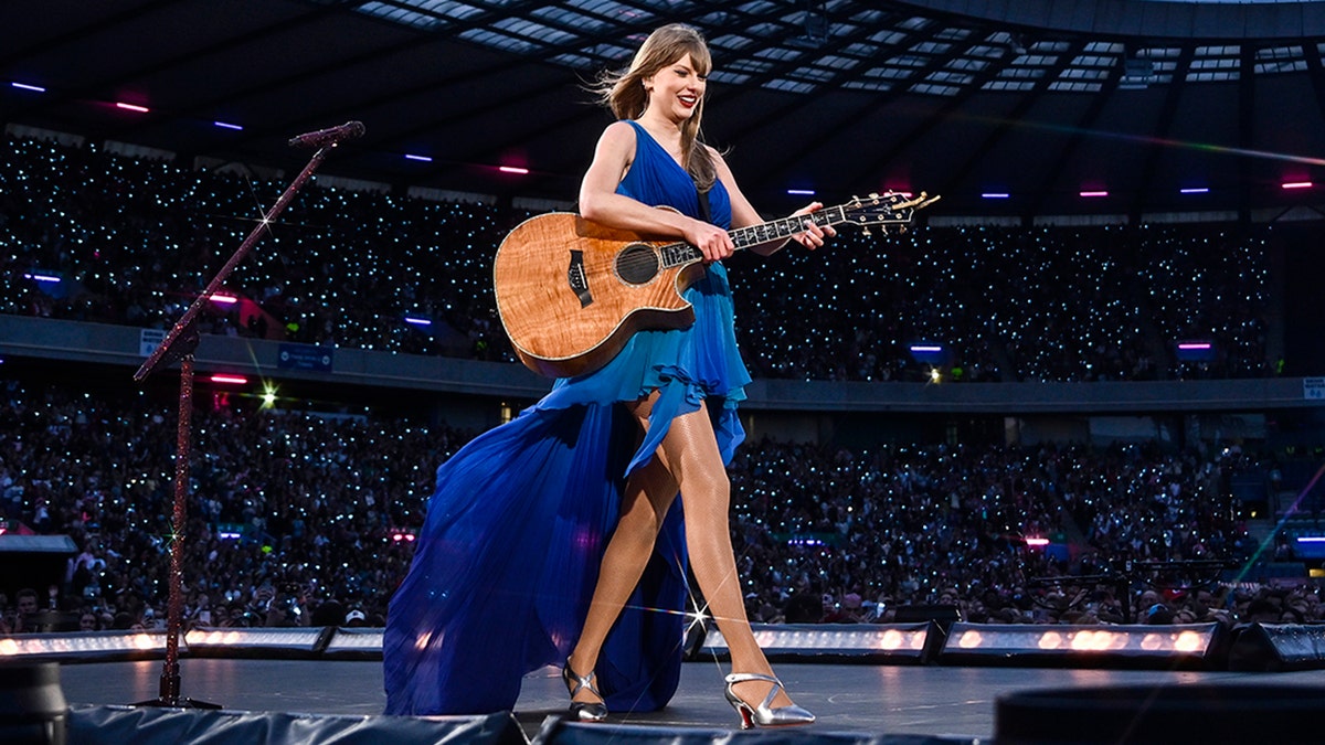 Taylor Swift in a long blue dress struts the stage while playing the guitar in Edinburgh 