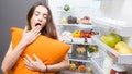 Young woman with pillow yawning standing in front of the refrigerator. Concept of eating during the night