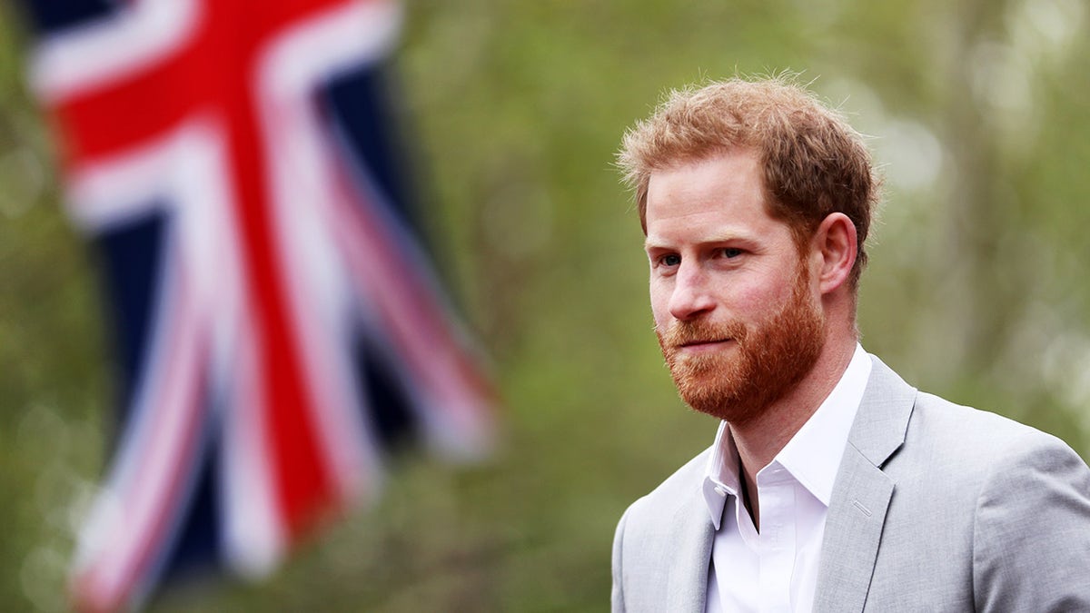 Prince Harry in a grey blazer and white shirt walking near a Union Jack flag.