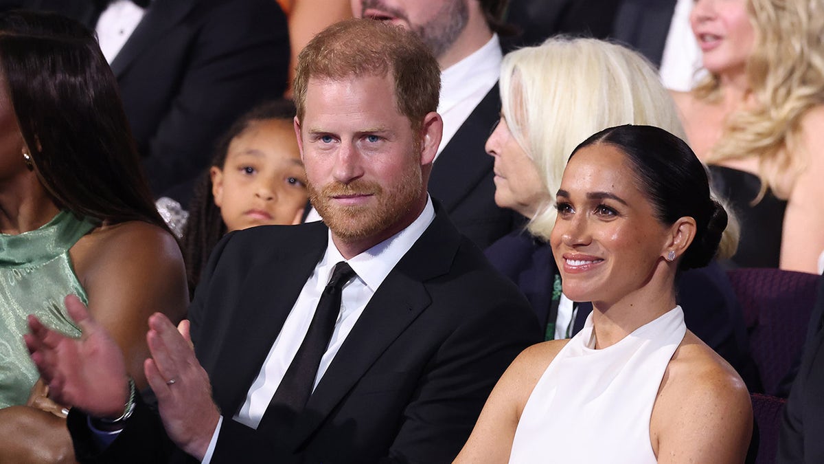 Meghan Markle in a white dress sitting next to Prince Harry in a black suit and tie.