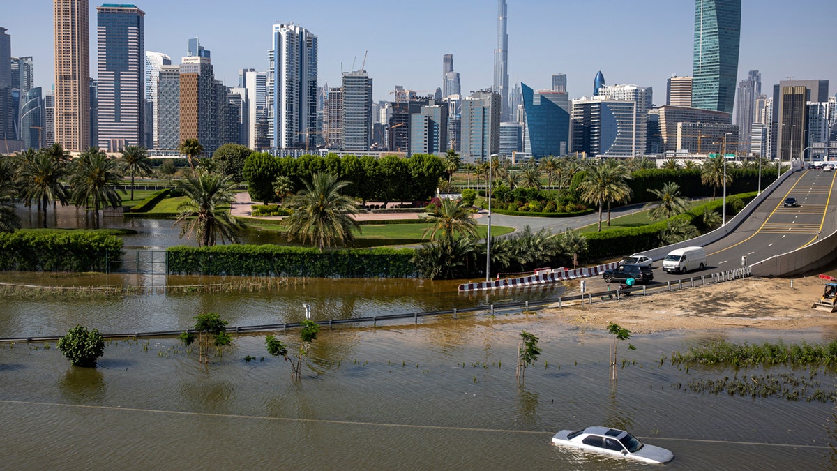 A major roadway in Dubai is seen flooded, with a half-submerged car in the foreground and the city skyline in the background.