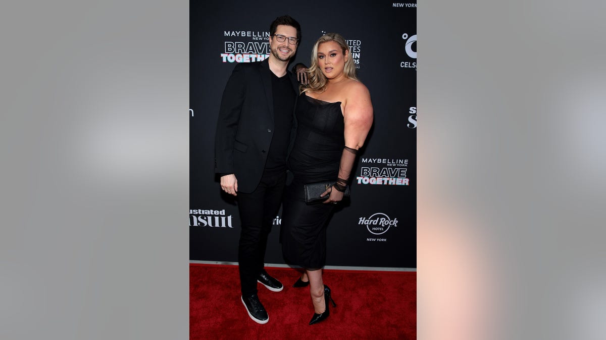 Hunter McGrady in a black dress posing next to her husband who is wearing black on the red carpet.