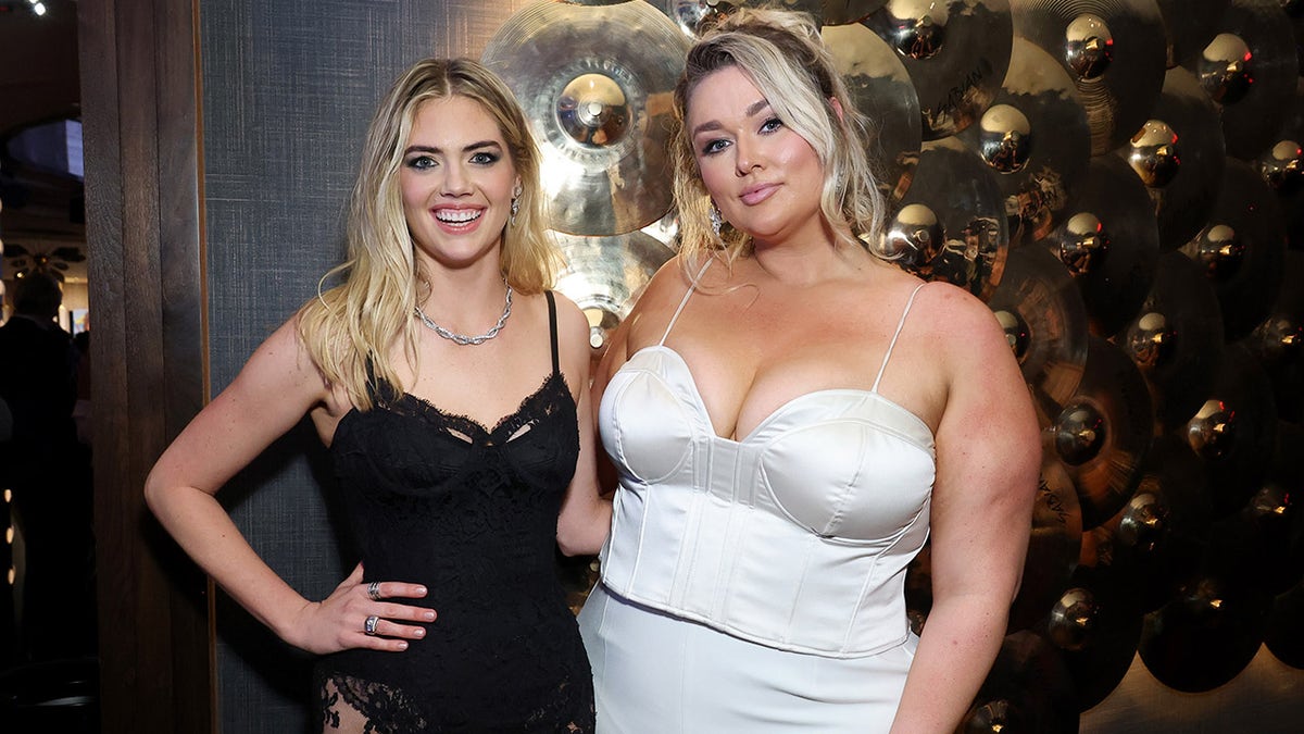 Kate Upton in a black dress posing next to Hunter McGrady in a white dress.