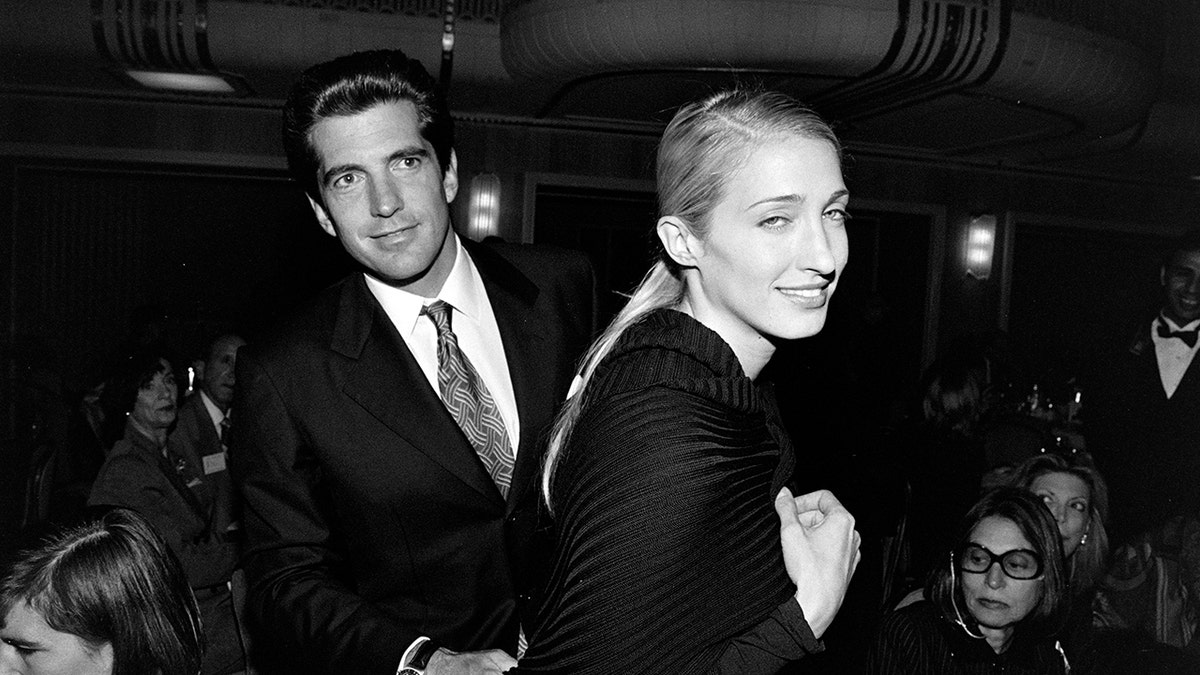 Carolyn Kennedy smiling at a photographer as John F. Kennedy Jr. looks away in a black and white photo.