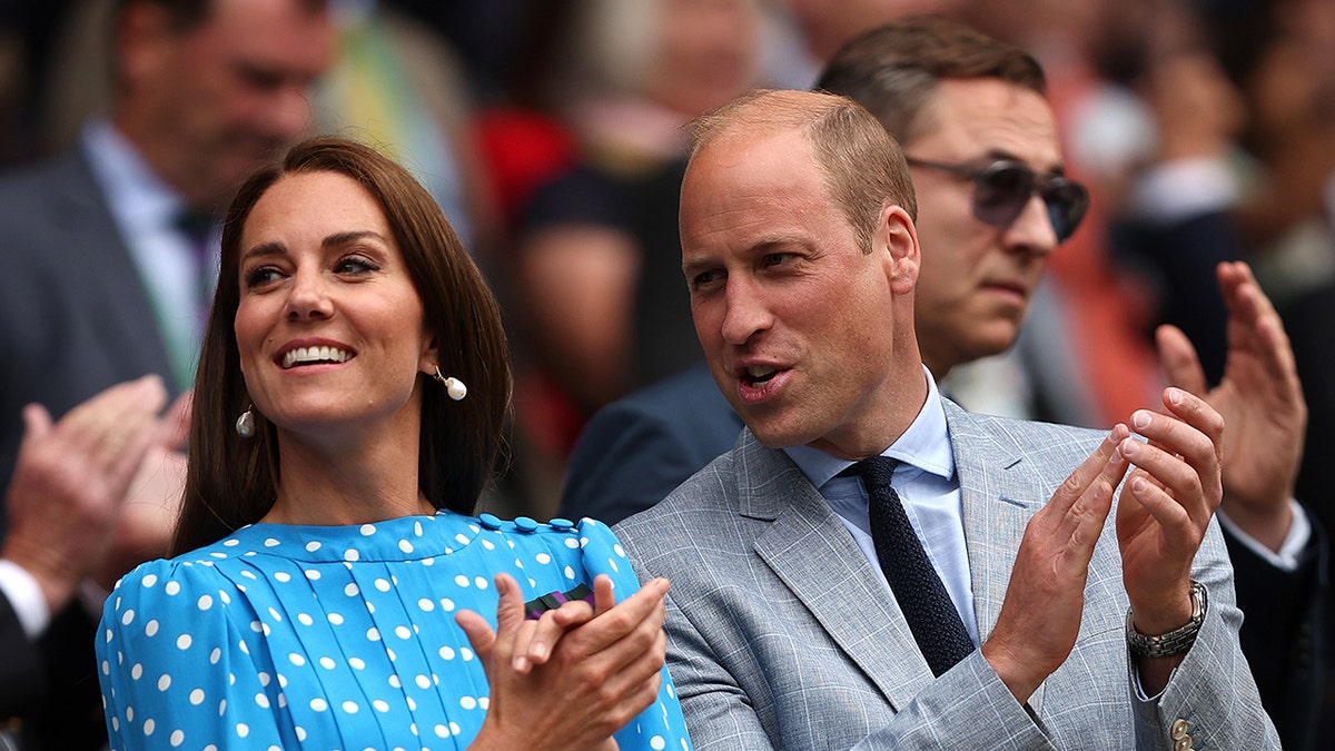 Prince William in a grey suit chatting to Kate Middleton from the stands as she wears a light blue and white polka dot dress.