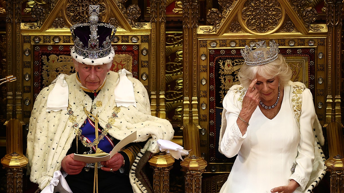 Queen Camilla scratches her head as King Charles reads from a pamphlet.