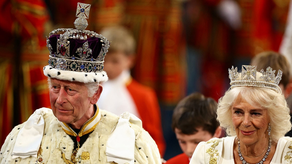 Queen Camilla walking next to King Charles as they both wear royal regalia and crowns.